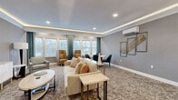 Located at 50 Mount Bethel Rd in Warren, NJ, this single-floor living room is a well-appointed space that combines refined design with practical functionality. The room features a tray ceiling with integrated ambient lighting that casts a warm, soft glow throughout, complemented by recessed downlights and stylish geometric pendant fixtures that add a decorative focal point above the wet bar area.

The wet bar is a standout architectural feature, complete with white cabinetry, a built-in sink, and a white countertop — ideal for entertaining. A bold black-framed glass door provides an elegant entryway, adding a contemporary contrast to the room's soft, neutral color palette.

Textured gray wallcovering lines the walls, lending depth and sophistication to the interior. Plush, patterned carpeting in warm earth tones anchors the space and adds comfort underfoot. Multiple seating arrangements — including a cushioned sofa, accent chairs, and a small dining area with dark chairs — allow for versatile use of the space.

A row of large windows fitted with white blinds and teal drapes floods the room with natural light and offers views of the surrounding wooded landscape. A wall-mounted split HVAC unit ensures year-round climate control. Framed wall art and carefully curated décor accessories complete the polished, upscale ambiance of this inviting living space.