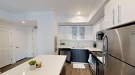 The kitchen showcases a contemporary design with a striking two-tone aesthetic, combining crisp white upper cabinetry with dark lower cabinets. Upper cabinets include glass-front display panels, adding visual depth while allowing for decorative storage. The countertops are finished in clean white, providing a bright, functional workspace throughout. A central island extends the prep area and serves as an additional surface for casual use, accented with small potted greenery for a touch of warmth. Stainless steel appliances, including a range, built-in microwave, dishwasher, and refrigerator, are seamlessly integrated into the layout, contributing to the kitchen's polished, modern character. The sink is positioned at the corner, maximizing efficiency within the thoughtfully arranged workspace. Recessed lighting in the tray ceiling provides consistent illumination, while wood-look flooring grounds the space with a warm, natural contrast to the otherwise cool palette. A doorway visible from the kitchen hints at an open, connected floor plan, enhancing the sense of flow throughout the single-level space.
