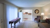 This single-floor residence offers a welcoming and functional layout designed for comfortable everyday living. The main living area is finished with soft wall-to-wall carpeting and neutral-toned walls, creating a warm and inviting atmosphere. Large windows allow natural light to filter into the space, enhancing the open feel of the room. A textured ceiling adds a classic architectural detail throughout.

The kitchen is positioned just off the living area and is clearly visible through an open passthrough, maintaining a sense of connectivity between the two spaces. It features white cabinetry extending to the upper walls, white appliances including a refrigerator and range, and light-colored countertops. A small accent rug adds a touch of warmth to the kitchen floor. Ample cabinet storage is provided both above and below the counters, making the kitchen highly practical.

The living area is spacious enough to accommodate a dining table and seating, demonstrating its versatility. A decorative round mirror adorns one wall, adding visual interest to the neutral palette. Overall, the space presents a clean, understated aesthetic with a layout that balances openness and defined functional zones.
