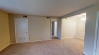 This single-floor residential space offers a clean and functional layout designed for comfortable everyday living. The main living area is finished with wall-to-wall carpeting in a warm neutral tone, providing a soft and inviting feel underfoot. The walls are painted in a light beige hue, creating a bright and open atmosphere that serves as a versatile backdrop for a variety of interior styles.

The space features standard white trim and baseboards that frame the rooms neatly, contributing to a cohesive and tidy aesthetic. Ventilation is well-distributed throughout, with ceiling vents visible in multiple areas to ensure consistent air circulation. A doorway connects the main living area to an adjacent hallway, suggesting additional rooms beyond and offering a sense of depth and connectivity within the floor plan.

A closet or storage area is accessible from the main room, providing practical organizational space. The layout flows naturally from one area to the next, making efficient use of the available square footage. The overall ambiance is understated and move-in ready, with neutral finishes that allow for easy personalization. This space is well-suited for those seeking a straightforward, low-maintenance living environment with a functional and adaptable interior design.