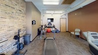 The fitness room is a thoughtfully designed exercise space that blends industrial character with practical functionality. A prominent exposed brick accent wall adds texture and visual interest, complementing the warm, earth-toned painted walls in contrasting shades of taupe and deep rust. Overhead, exposed ductwork and fluorescent lighting run across the ceiling, reinforcing the room's industrial aesthetic while providing ample illumination throughout.

The space is well-stocked with a variety of cardio equipment, including treadmills, an elliptical trainer, a stationary bike, and a stair climber, offering users multiple options for cardiovascular exercise. A weight bench with barbells and free weights supports strength training needs. A dedicated functional training area includes an exercise ball, step platform, resistance accessories, and yoga mats, providing space for flexibility and bodyweight workouts.

A wall-mounted flat-screen television adds convenience for entertainment during workouts. The carpeted flooring with rubber mat sections beneath equipment ensures both comfort and safety. Natural light enters through a window fitted with a roller shade, while a water dispenser and waste bin contribute to the room's overall practicality. Seating is available along the perimeter for rest between exercises.