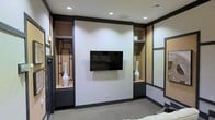Located at 75 Cross Creek Pkwy in Hattiesburg, Mississippi, this single-story space presents a polished and contemporary interior designed with both style and function in mind. The space showcases a refined aesthetic, highlighted by a striking feature wall that serves as a central focal point. A wall-mounted flat-screen display is flanked by symmetrical built-in shelving units, framed in dark trim with geometric lattice detailing that adds a modern, architectural flair. The open shelving niches provide display space accented by warm tan backing, while enclosed cabinetry below offers practical storage. The overall color palette balances crisp white walls with bold dark trim and baseboards, creating a clean and professional contrast throughout. Recessed ceiling lighting provides even, well-distributed illumination across the interior, contributing to a bright and welcoming atmosphere. The flooring features neutral-toned carpet, adding warmth underfoot while maintaining a cohesive, understated look. Mirrored wall panels further enhance the sense of openness, reflecting light and adding depth to the interior. The overall ambiance is modern, orderly, and thoughtfully appointed, making this space well-suited for professional or commercial use.