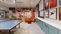 Located at 1501 Euclid Ave in Cleveland, Ohio, this dynamic game room delivers an engaging and visually stimulating environment designed for recreation and socializing. The space is thoughtfully divided into distinct activity zones, each with its own character and energy.

At one end, a vibrant blue accent wall sets a lively backdrop for a classic billiards table, complemented by a wall-mounted flat-screen TV and a modern circular pendant light. Comfortable seating is tucked nearby, offering a relaxed spot to watch the action unfold.

The opposite zone features a full ping-pong table positioned beneath a row of sleek black pendant lights. This area is anchored by a striking large-scale abstract mural that spans the full wall, bursting with bold reds, blues, and earth tones. A cluster of plush orange barrel chairs and rounded ottomans in warm terracotta tones creates an inviting lounge area in front of the mural, ideal for casual conversation or downtime between games.

Teal-painted cabinetry with paneled doors lines one wall, providing built-in storage while adding a refined, polished touch. Broadway-themed and artistic framed posters add a culturally inspired layer of décor throughout. The flooring features a neutral geometric-patterned tile that ties the space together cohesively. Recessed lighting throughout ensures the entire room is well-lit and welcoming.