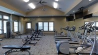 This fitness center is designed to support a well-rounded workout experience within a bright, spacious environment. The room features striking vaulted ceilings that amplify the sense of openness, complemented by ceiling fans and a combination of skylight-style and recessed lighting that keeps the space well-lit throughout the day. Natural light also filters in through a set of windows along the back wall, adding a welcoming ambiance to the room.

The facility is outfitted with a comprehensive selection of cardio equipment, including multiple treadmills, stationary upright bikes, and cycling bikes arranged to allow comfortable spacing between users. Strength and resistance training equipment is also available, positioned toward the rear of the room for a defined workout zone. A wall-mounted television is installed at the front of the space, providing entertainment during workouts.

The flooring consists of durable, patterned carpet tiles suited for a fitness environment, and the neutral tan and gray wall tones give the space a clean, professional appearance. A dedicated entry door provides direct access to the facility. Overall, the fitness center offers a functional and comfortable setting for a variety of exercise routines.
