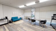 Located at 170 E Griffith St in Jackson, Mississippi, this single-floor multipurpose space offers a well-organized and versatile layout designed to serve a variety of functional needs. The interior features light wood-style flooring throughout, giving the space a clean and cohesive aesthetic.

The main open area serves as both a lounge and a meeting space. A vibrant teal sofa anchors the lounge section, set atop a round area rug and accompanied by motivational wall art and a side table with a lamp. Adjacent to the lounge, a modern gray conference table with contemporary molded chairs provides a dedicated area for meetings or collaborative work. Large windows fitted with horizontal blinds allow natural light to fill the room while maintaining privacy.

A separate area features built-in multi-tier shelving along one wall, ideal for storage or package management, alongside a wall-mounted flat-screen television. A door from this area leads to a room with gold-tone mailboxes, suggesting a shared or communal building environment.

Fitness equipment, including a stationary bike, is also present within the space, adding to its multi-use character. A small appliance station with a microwave and coffee maker provides basic kitchen convenience. Exposed conduit along the ceiling contributes an industrial design accent throughout the space.
