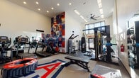 Located at 255 N Granada Ave in Tucson, Arizona, this single-floor fitness center is a dynamic and fully equipped workout facility designed to inspire and accommodate a wide range of training styles. The space features soaring ceilings that lend an open, airy feel, enhanced by recessed lighting, skylights, and a ceiling fan that keep the environment bright and comfortable. A bold, eye-catching collage mural in red, white, and blue anchors one wall, while a large University of Arizona-themed area rug with the iconic "A" logo adds strong school spirit to the floor. Cardio equipment including treadmills, ellipticals, and stationary bikes lines one side of the room, all positioned in front of wall-mounted mirrors that amplify the sense of space. The strength training area is equally well-appointed, featuring a multi-functional rack stocked with medicine balls and barbells, a heavy punching bag, adjustable benches, plyo boxes, and a large tire for functional training. A wall-mounted TV provides entertainment, while large glass doors open directly to an outdoor pool area, flooding the space with natural light and offering refreshing views. The "Find Your Fit" signage reinforces the motivating atmosphere throughout.