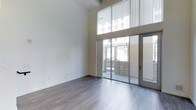 This thoughtfully designed two-floor space makes a striking first impression with its dramatic double-height ceilings and expansive floor-to-ceiling windows that flood the interior with abundant natural light. The main living area showcases clean, contemporary architecture with smooth white walls that enhance the sense of openness and volume throughout. Large sliding glass doors open directly to an outdoor area, seamlessly blending indoor and outdoor living while providing easy access to fresh air and exterior views. The wide-plank, hardwood-style flooring in a soft gray tone runs throughout, adding warmth and cohesion to the space while complementing the bright, neutral color palette. Vertical blinds on the upper windows offer flexible light control without compromising the airy feel. The minimalist design aesthetic is consistent throughout, with architectural details kept intentionally clean and unobtrusive, allowing the scale and natural light to take center stage. The two-floor layout provides distinct zones for living and additional functional use, offering versatility to suit a variety of needs. Overall, the space exudes a modern, refined ambiance that is both functional and visually compelling.