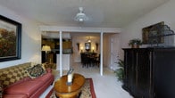 This single-story residence showcases an open-concept floor plan that seamlessly connects the living room, dining area, and kitchen, creating a cohesive and functional living space. The living room serves as a welcoming focal point, featuring light-toned carpeting, a ceiling fan for year-round comfort, and a textured ceiling. A warm, oval wooden coffee table anchors the seating area, complemented by a red upholstered sofa and a patterned area rug that adds visual interest to the space. A dark wood armoire provides both storage and a striking contrast against the neutral walls.

Structural columns define the transition between the living and dining areas without interrupting the open flow. The dining area accommodates a full dining set with dark wood chairs, and a pendant light fixture overhead adds an elegant touch. Decorative plants and wall sconces contribute to the warm ambiance throughout.

The kitchen, visible beyond the dining area, features warm wood cabinetry and a raised breakfast bar that separates it from the main living space, offering additional seating and serving functionality. A six-panel door leads to additional areas of the home, completing this well-appointed, single-level layout.