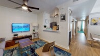 This well-appointed single-floor residence offers a thoughtfully designed layout that balances comfort and modern style. The open-concept living area serves as the heart of the home, featuring warm hardwood-style flooring, a ceiling fan, and a wall-mounted television. A vibrant abstract artwork anchors the living room, complemented by contemporary furniture and a bold blue-toned area rug that ties the space together.

The galley-style kitchen is both functional and visually striking, with two-tone cabinetry featuring dark lower cabinets and white uppers, stainless steel appliances including a refrigerator, range, and microwave, plus a convenient pass-through shelf that connects to the living area. A dedicated dining area accommodates a round glass-top table with modern white chairs, bathed in natural light from a large window dressed with teal curtains.

Two bedrooms offer restful retreats, each with hardwood-style flooring, ample natural light, and coordinated teal accents. The primary bathroom includes a vessel sink vanity with a bold round mirror and dark hardware, while a second full bathroom features a tub-shower combination. A consistent palette of whites, teals, and warm wood tones unifies the entire space, creating a cohesive and inviting atmosphere throughout.
