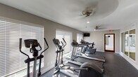 Located at 4010 Linkwood Dr in Houston, Texas, this fitness center is a single-floor facility designed to support a comprehensive workout experience. The space is thoughtfully laid out to accommodate a variety of exercise modalities, from cardiovascular training to strength work.

The main cardio area is lined with large windows dressed in white blinds, allowing natural light to fill the room and create an open, energizing atmosphere. The room is equipped with multiple treadmills, elliptical trainers, and a stationary bike, all from Life Fitness. Ceiling fans and recessed lighting provide comfort and visibility throughout. A wall-mounted flat-screen television is positioned for easy viewing during workouts.

A dedicated strength training alcove houses a robust dual-cable functional trainer, ideal for a wide range of resistance exercises. The durable, dark speckled carpet flooring throughout the space provides cushioning and stability underfoot.

Additional amenities include a stability ball storage rack holding multiple exercise balls, and a built-in water fountain station conveniently accessible along the interior corridor. An emergency exit door with exterior access adds practicality to the layout. The neutral gray walls and clean white trim give the facility a modern, professional appearance that is both functional and welcoming.