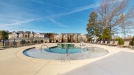 Located at 708 Glamorgan Way in Rock Hill, South Carolina, this outdoor amenity space serves as the centerpiece of a well-appointed residential community. The focal point is a generously sized resort-style swimming pool with a curved, freeform shape, complemented by a separate shallow wading area — ideal for relaxing on warm Southern days. The expansive concrete sun deck surrounds the pool and is lined with multiple lounge chairs, providing ample space for sunbathing and outdoor leisure. The pool area is enclosed by a sturdy black metal fence, ensuring both safety and privacy, with a dedicated gated entry point for controlled access. A charming pool house anchors the space, featuring a covered porch with a cupola roofline, shiplap-style siding, and tasteful landscaping including upright evergreen shrubs. The structure adds architectural character while providing functional support amenities for pool visitors. Mature trees frame the perimeter of the space, offering natural shade and a sense of seclusion. Views of the surrounding multi-story residential buildings are visible in the background, reinforcing the community setting. Under bright, open skies, this thoughtfully designed outdoor space offers a welcoming and well-maintained retreat for residents throughout the warmer months.