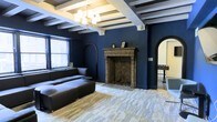 This captivating single-floor space makes an immediate impression with its deep navy blue walls, setting a bold and dramatic tone throughout. The ceiling draws the eye upward with a series of exposed white-painted structural beams arranged in a coffered pattern, lending a sense of architectural depth and character. A classic chandelier with warm-toned bulbs hangs centrally, providing elegant ambient lighting that complements the natural light filtering in through the windows.

The focal point of the main living area is an ornate, intricately carved stone fireplace, featuring detailed craftsmanship and a herringbone-patterned firebox interior. This statement piece anchors the room and adds a sense of historic grandeur. The living area is comfortably appointed with a large, low-profile sectional sofa in dark leather, resting atop light-toned hardwood floors that provide a striking contrast against the dark walls.

Graceful arched doorways frame the entrances to adjoining spaces, adding a refined architectural touch that carries consistently through the design. A glimpse through one arch reveals an additional recreation area, hinting at the space's versatility. The overall ambiance is one of moody sophistication, balancing classic architectural details with a contemporary, curated aesthetic.