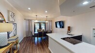 This well-appointed single-floor space showcases an open-concept layout that seamlessly connects the kitchen, dining, and living areas. Rich, dark hardwood flooring runs throughout, lending warmth and continuity to the space.

The kitchen features a large island with a crisp white quartz countertop, an electric cooktop, and an undermount sink. Dark cabinetry on the island base provides a striking contrast to the bright surfaces above. A stainless steel refrigerator and recessed lighting complete the functional yet refined kitchen design.

The dining area is a standout feature, centered around a substantial wood dining table with upholstered chairs. Overhead, two matching geometric lantern-style pendant lights add an elegant, modern-farmhouse aesthetic. Black-framed French doors with decorative ironwork draw the eye and open to an outdoor area, flanked by floor-length linen drapes that soften the space. A wall-mounted flat-screen television is positioned nearby for added versatility.

A console table styled with a round mirror, decorative lamp, and curated artwork creates a welcoming focal point near the entry. The front door, also black-framed with glass panel accents, introduces natural light from the entryway. Crown molding and recessed lighting throughout add polished architectural detail, contributing to the space's cohesive and sophisticated ambiance.