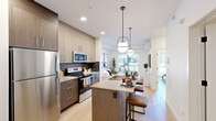 Located in Calgary's Hartell Way SE, this thoughtfully designed one-bedroom, one-bathroom unit makes excellent use of its compact footprint across a single floor. The open-concept layout connects the kitchen and living room seamlessly, creating a bright and functional living space.

The kitchen is well-equipped with stainless steel appliances, including a full-size refrigerator, electric range, and over-the-range microwave. Two-tone cabinetry — combining warm wood-grain lower cabinets with light upper cabinets — pairs with a dark tile backsplash and light quartz countertops, lending a contemporary aesthetic. Light hardwood-style flooring runs throughout the unit, adding warmth and continuity.

The living room is anchored by a large sliding glass door that opens onto a private patio, offering views of the landscaped courtyard and building exterior. Natural light fills the space, creating an airy atmosphere.

The bedroom features a bright window overlooking the greenery outside and includes a closet with sliding door access. A dedicated office nook provides a quiet, functional workspace. The bathroom is finished with a clean, all-white tub-shower combination.

The building exterior showcases attractive landscaping, open green space, and an outdoor sports court, offering residents well-rounded amenity access.
