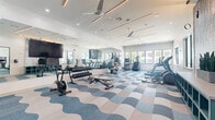 Located at 1800 Central Pine St in Montgomery, Texas, this fitness center is a thoughtfully designed, single-floor facility that combines functionality with contemporary style. The space features a striking wave-patterned carpet in blue and cream tones that adds a dynamic visual element throughout the room, complementing the bright, white walls and modern aesthetic.

The facility is generously proportioned, offering distinct zones for various types of exercise. The cardio area is equipped with multiple treadmills, elliptical trainers, stair climbers, and stationary bikes, all positioned near large windows that provide abundant natural light and views of the surrounding community. A dedicated free weights section features a full dumbbell rack, adjustable benches, and a cable machine for strength training. Floor-to-ceiling mirrors along one wall enhance the sense of space and allow users to monitor their form during workouts.

Overhead, linear accent lighting and ceiling fans with recessed lighting create a clean, well-lit environment. A large flat-screen TV is mounted prominently for entertainment. A decorative accent wall featuring an abstract, nature-inspired mural adds an artistic focal point to the space. The facility also includes a teal storage credenza for organizational use. The overall ambiance is bright, energizing, and modern, making it a well-rounded fitness destination.