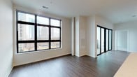 This contemporary single-floor unit offers a clean, modern aesthetic throughout, combining thoughtful design with functional living spaces. The open living area is anchored by large, black-framed floor-to-ceiling windows that frame sweeping city skyline views, flooding the space with natural light. Rich, dark hardwood flooring runs throughout the main living and kitchen areas, adding warmth to the bright white walls.

The kitchen is well-appointed with a full suite of stainless steel appliances, including a French door refrigerator, range, built-in microwave, and dishwasher. Crisp white cabinetry extends to the ceiling for ample storage, complemented by a classic white subway tile backsplash and a quartz waterfall-edge island, perfect for dining or meal preparation.

The bedrooms feature plush neutral carpeting and generously sized black-framed windows that continue the city views. One bedroom connects to a walk-in closet equipped with multi-tiered wire shelving systems. Two bathrooms serve the unit: one features a sleek sliding glass shower enclosure, navy cabinetry, and large-format tile flooring, while the second offers a dark vanity with a backlit mirror and matching tile work. The overall ambiance is clean, contemporary, and urban.