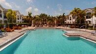 Located at 11496 Sumter Grove Circle in Naples, Florida, this outdoor amenity space offers a resort-inspired pool experience within a well-appointed residential community. The centerpiece of the space is a generously sized, crystal-clear swimming pool with striking blue tile accents, complemented by a second pool area that together create an expansive aquatic retreat. Both pools are surrounded by wide, paver-finished sun decks that provide ample room for lounging and recreation under the Florida sun.

The pool deck is furnished with an array of cushioned lounge chairs and chaise seating, accented with vibrant red and blue cushions that add a lively pop of color to the setting. Private cabanas with draped curtains are positioned along one side of the larger pool, offering shaded retreats for relaxation. Lush tropical landscaping, including towering palm trees, manicured hedges, and colorful plantings, frames the entire area, reinforcing the resort-like ambiance.

Surrounding the pool areas, the community's multi-story Mediterranean-style buildings with white stucco exteriors, arched balconies, and tile rooflines create an elegant architectural backdrop. A dedicated clubhouse structure with covered outdoor seating is also visible, adding to the functional appeal of the space. The entire outdoor area is enclosed by a wrought iron fence, ensuring privacy and security throughout.