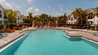 Located at 11496 Sumter Grove Circle in Naples, Florida, this outdoor amenity space offers a resort-inspired pool experience within a well-appointed residential community. The centerpiece of the space is a generously sized, crystal-clear swimming pool with striking blue tile accents, complemented by a second pool area that together create an expansive aquatic retreat. Both pools are surrounded by wide, paver-finished sun decks that provide ample room for lounging and recreation under the Florida sun.

The pool deck is furnished with an array of cushioned lounge chairs and chaise seating, accented with vibrant red and blue cushions that add a lively pop of color to the setting. Private cabanas with draped curtains are positioned along one side of the larger pool, offering shaded retreats for relaxation. Lush tropical landscaping, including towering palm trees, manicured hedges, and colorful plantings, frames the entire area, reinforcing the resort-like ambiance.

Surrounding the pool areas, the community's multi-story Mediterranean-style buildings with white stucco exteriors, arched balconies, and tile rooflines create an elegant architectural backdrop. A dedicated clubhouse structure with covered outdoor seating is also visible, adding to the functional appeal of the space. The entire outdoor area is enclosed by a wrought iron fence, ensuring privacy and security throughout.