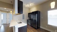 This single-floor space showcases a contemporary kitchen designed with both style and functionality in mind. The kitchen is equipped with a full suite of stainless steel appliances, including a French door refrigerator with bottom freezer, a stainless electric range, and a built-in microwave, all complemented by dark espresso cabinetry that creates a bold contrast against the crisp white countertops. A stainless steel undermount sink with a modern arc-neck faucet anchors the counter space, while a sleek wall-mounted range hood adds an industrial design element overhead. Track lighting installed on the ceiling provides focused, adjustable illumination throughout the cooking area. The rich, dark hardwood flooring runs throughout the space, adding warmth and depth to the overall aesthetic. Neutral-toned walls contribute to a clean, open atmosphere, and generous cabinetry offers ample storage. The thoughtful layout balances practicality with modern design, making the kitchen both an efficient workspace and a visually appealing centerpiece of the unit.