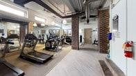 The Depot Fitness Center combines industrial architectural character with a comprehensive range of workout equipment, creating a functional and visually distinctive exercise environment. Exposed ductwork and structural ceiling elements lend an authentic industrial aesthetic, while pendant lighting and ceiling fans maintain a comfortable, well-lit atmosphere throughout the space.

The main exercise area features a robust selection of cardio equipment, including treadmills, elliptical trainers, a stationary bike, and a stair climber, all arranged across rubber-matted flooring. Full-length mirrors line the walls, amplifying the sense of space and allowing users to monitor their form during workouts. High clerestory windows allow natural light to filter in, enhancing the open feel of the room.

A dedicated strength training zone houses a varied collection of kettlebells, dumbbells, weight benches, and a Paramount multi-station cable machine, providing ample options for resistance training. Exposed brick columns add architectural warmth and serve as defining structural elements between zones.

The entry area features light wood-tone flooring, visible HVAC systems overhead, and direct elevator access, ensuring convenient entry. The overall ambiance is utilitarian yet inviting, balancing the building's historic character with modern fitness functionality.