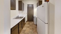 Located at 1100 E Liberty St in Girard, Ohio, this single-floor unit offers a straightforward and functional layout across several distinct spaces. The living area is generously proportioned, featuring neutral-toned carpet, cream-colored walls, dark baseboard trim, and a wall-mounted air conditioning unit alongside baseboard heating. A window with blinds admits natural light, contributing to a bright and open feel. A hallway connects the main living space to the private areas of the unit, maintaining a clear and practical flow throughout.

The bedrooms are carpeted and finished with the same neutral palette, each offering adequate space for furnishings. One bedroom includes a built-in closet with a sliding door and shelving, providing organized storage. Additional rooms with tile flooring offer flexible utility space suitable for various purposes.

The galley-style kitchen is equipped with dark cabinetry, white countertops, a stainless steel sink, and includes a refrigerator and range. Tile flooring completes the kitchen area, which also features a door providing secondary access. The bathroom includes a full-size tub and shower combination, toilet, and tile flooring, presenting a clean and functional design. Overall, the unit maintains a consistent, no-frills aesthetic throughout.