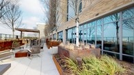 This elevated outdoor terrace is defined by its modern architectural design and thoughtfully arranged amenities. The building's exterior showcases a warm-toned brick facade in varying shades of cream and tan, providing a refined backdrop to the open-air space. Floor-to-ceiling glass curtain walls line portions of the terrace, reflecting the surrounding skyline and allowing natural light to flow between indoor and outdoor areas.

The terrace is organized into distinct functional zones. One section features a glass-panel railing system mounted atop a textured brick half-wall, offering unobstructed panoramic views of the urban landscape and open roadways below. Plush neutral-toned lounge chairs are positioned here to take full advantage of the elevated vistas. Adjacent areas incorporate sleek, low-profile sofas set on composite wood decking beside large Corten steel planters, which contain ornamental grasses and deciduous trees that add organic texture throughout the space.

A separate dining and bar area offers high-top tables and chairs beneath a covered pergola structure, ideal for gatherings. Continuous large-format tile pavers unify the various zones, maintaining a cohesive, polished aesthetic throughout. The overall ambiance is contemporary and open, with a strong connection to the surrounding cityscape.
