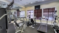 This fitness room offers a focused environment for cardiovascular and strength training. The space is outfitted with a variety of cardio equipment, including treadmills, an elliptical trainer, a stationary recumbent bike, and additional exercise machines, providing users with diverse workout options. A large wall-mounted flat-screen television is positioned prominently above the windows, allowing residents to stay entertained during their workouts. The flooring consists of durable dark rubberized matting, ideal for high-traffic fitness use. Multiple windows dressed with rich wood-toned horizontal blinds line the walls, allowing natural light to filter into the room while maintaining privacy. A full-length mirror along one wall visually expands the space and assists with exercise form. The room features classic crown molding and a neutral warm-toned wall color, lending a polished, refined feel. Recessed lighting in the ceiling ensures the space remains well-lit at all times. The overall ambiance is clean, practical, and inviting, making it a functional amenity well-suited for daily fitness routines.