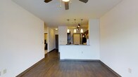 This single-floor residence features an open-concept layout that integrates the living and kitchen areas into one cohesive, functional space. Rich, dark hardwood-style flooring runs throughout, providing a warm contrast against the clean white walls and creating a polished, contemporary aesthetic.

The living area is anchored by a ceiling fan overhead, offering both comfort and airflow. The space flows naturally toward a kitchen peninsula, which serves as both a workspace and an informal dining counter. The peninsula is topped with light granite-style countertops, providing a bright counterpoint to the dark cabinetry. Above the peninsula, a cluster of sleek pendant lights hang at varying heights alongside a track lighting system, ensuring the kitchen is both stylish and well-illuminated.

The kitchen is equipped with stainless steel appliances and dark espresso-toned cabinetry, reinforcing the modern design language throughout. A hallway visible from the main living area leads to additional rooms, hinting at a well-organized floor plan. The overall ambiance is modern and clean, with thoughtful lighting and consistent finishes lending the space a cohesive, move-in-ready appeal.