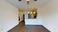 This single-floor residence features an open-concept layout that integrates the living and kitchen areas into one cohesive, functional space. Rich, dark hardwood-style flooring runs throughout, providing a warm contrast against the clean white walls and creating a polished, contemporary aesthetic.

The living area is anchored by a ceiling fan overhead, offering both comfort and airflow. The space flows naturally toward a kitchen peninsula, which serves as both a workspace and an informal dining counter. The peninsula is topped with light granite-style countertops, providing a bright counterpoint to the dark cabinetry. Above the peninsula, a cluster of sleek pendant lights hang at varying heights alongside a track lighting system, ensuring the kitchen is both stylish and well-illuminated.

The kitchen is equipped with stainless steel appliances and dark espresso-toned cabinetry, reinforcing the modern design language throughout. A hallway visible from the main living area leads to additional rooms, hinting at a well-organized floor plan. The overall ambiance is modern and clean, with thoughtful lighting and consistent finishes lending the space a cohesive, move-in-ready appeal.