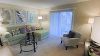 This single-floor residential space offers a welcoming and well-organized layout designed for comfortable everyday living. The living room serves as the heart of the home, featuring soft wall-to-wall carpeting and warm, neutral-toned walls that create a calm and inviting atmosphere. A large sliding glass door dressed with vertical blinds fills the room with abundant natural light while providing direct access to an outdoor area, enhancing the sense of openness and connectivity to the exterior. The living area offers ample room for a generously sized sectional sofa and additional seating, making it ideal for both relaxing and entertaining. Decorative wall art adds personality and a touch of style to the space. A glass-top coffee table anchors the seating arrangement, complementing the overall light and airy aesthetic. The layout flows naturally and efficiently, reflecting a practical yet thoughtfully designed living environment. Overall, the space presents a clean, functional, and comfortable residential setting with key features including natural light, carpeted flooring, and convenient outdoor access.