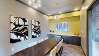 Located in Frisco, this single-floor space is a beautifully designed conference and dining room that blends modern aesthetics with functional elegance. The room is anchored by a long, rich dark-wood conference table surrounded by cream upholstered chairs, offering ample seating for meetings or private dining events. Two dramatic sputnik-style chandeliers serve as eye-catching focal points, casting a warm, ambient glow throughout the space. The walls feature a sophisticated two-tone color scheme — soft gray paired with a bold, energizing yellow accent wall — creating a dynamic yet cohesive visual atmosphere. Large-scale abstract artwork with bold brushstrokes in black, gold, and blue adds a curated, gallery-like quality to the room. A sleek built-in credenza in dark charcoal lines the accent wall beneath a framed art piece, offering both storage and display surfaces. The tray ceiling with recessed lighting adds architectural depth, while wood-look flooring grounds the space with warmth and texture. Overall, this space exudes a contemporary, professional ambiance suited for collaborative work sessions, private events, or executive gatherings.