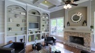 This single-floor space makes a strong impression with its thoughtfully designed living area, where elegance and functionality come together seamlessly. The focal point of the room is a floor-to-ceiling built-in shelving unit featuring arched display niches, beadboard backing, and a centrally mounted flat-screen television — offering both stylish storage and a natural entertainment hub. The warm hardwood flooring grounds the space, while the coffered ceiling with a ceiling fan adds architectural depth and visual interest overhead. A striking stone fireplace anchors one wall, providing a rustic yet refined contrast to the room's clean, neutral color palette of soft grays and whites. Large windows invite generous natural light into the space and frame views of lush greenery and an outdoor area beyond, creating a bright and airy ambiance. Contemporary black leather seating and glass-topped accent tables furnish the room with a modern edge. The combination of classic architectural details — from the arched built-ins to the tray ceiling — with modern furnishings gives this space a timeless, welcoming character well-suited for both relaxing and entertaining.