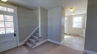 This two-floor space offers a straightforward and functional layout defined by clean lines and neutral tones. The main level features a combination of carpet and tile flooring, transitioning between the living area and the entryway. The carpeted section provides a comfortable feel underfoot, while the tiled entryway near the front door adds durability to the high-traffic zone. A carpeted staircase with white-painted risers and trim connects the two floors, anchored by crisp white baseboards that run throughout the space, lending a tidy, cohesive appearance. The walls are finished in a soft, muted gray-beige palette, creating a calm and neutral atmosphere. The entryway benefits from natural light streaming through a window adjacent to the front door, brightening the space and creating an inviting first impression. Ceiling-mounted flush light fixtures provide additional illumination across both the living area and the entry zone. A baseboard heating unit is visible along the wall near the entryway, indicating a practical heating solution. Overall, the space presents a clean, move-in-ready environment with a modest and understated aesthetic suited to a variety of uses.