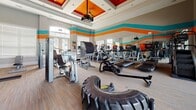 Located at 4601 Monroe Way in Fredericksburg, Virginia, this single-level fitness center is designed to deliver a comprehensive workout experience within a vibrant, energizing environment. The space is immediately distinguished by bold horizontal stripes of teal, orange, and gray that wrap around the walls, lending a dynamic, modern aesthetic throughout.

The facility is generously sized, accommodating a wide array of equipment across distinct workout zones. A dedicated cardio section features Matrix-brand treadmills, stair climbers, ellipticals, and rowing machines arranged in a row along a mirrored wall, maximizing the sense of openness. A large flat-screen projector is mounted above, adding an entertainment element to cardio sessions.

The strength training area is equally well-appointed, with multiple cable and selectorized machines, a rack of dumbbells, TRX suspension trainers, a medicine ball station, and a heavy tractor tire for functional training. The coffered ceiling with a ceiling fan adds architectural character overhead.

Natural light pours in through floor-to-ceiling glass doors and large windows. Sliding glass doors on one side open directly to a resort-style outdoor pool and courtyard, while French doors on the opposite end face the surrounding community, keeping the space bright and well-ventilated throughout the day.