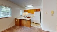 Located at 1424 Pacific Ave in Natrona Heights, Pennsylvania, this single-floor unit offers a practical and well-organized layout. The living area is finished with warm neutral carpet and features a ceiling fan with light fixture, a textured ceiling, and a window dressed with blinds that allows natural light to fill the space. An open passageway connects the living area to the kitchen, which is outfitted with warm-toned wood cabinetry, a white appliance suite including a range, dishwasher, and refrigerator, and a peninsula with a light countertop that provides additional prep and casual dining space. The kitchen floor features tile-style flooring, clearly delineating it from the carpeted living zones. The bedroom is a quiet, carpeted room with two windows fitted with blinds, offering ample natural light and a comfortable, neutral atmosphere. The full bathroom is clean and functional, featuring a tub and shower combination with panel surround, a vanity with a wood cabinet base and white countertop sink, and windows with frosted glass for privacy. The overall layout, as visible in the dollhouse view, is efficiently organized with well-defined spaces flowing from the kitchen and living area through to the bedroom and bathroom.
