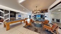 This single-floor amenity space is thoughtfully designed to blend social, recreational, and lounge functions within a cohesive contemporary aesthetic. The layout flows across several distinct zones, each finished with care and attention to detail.

The main lounge area features large-format light tile flooring, a statement chandelier with globe bulbs, and an open shelving unit displaying books and décor. Cognac leather chairs surround a textured stone coffee table, creating an inviting gathering spot. Adjacent to this area, a full-size billiards table anchors a more active zone beneath recessed lighting and a coffered ceiling detail.

A cozy lounge nook offers a sectional leather sofa, a dark ceiling treatment, and an eye-catching arrangement of circular gold-framed mirrors mounted against a soft gray wallcovering. A sculptural chevron-patterned wall serves as a dramatic divider between zones.

A separate lounge features a wall-mounted TV above a linear fireplace set into a marble-clad accent wall, with plush seating arranged for relaxed viewing. The kitchenette includes a coffee station, microwave, marble backsplash, and warm wood cabinetry. A bar counter with pendant lighting and exterior-facing windows provides a bright café-style seating area. The lobby entrance features floor-to-ceiling glass partitions and a stone-clad elevator surround. A golf simulator completes the recreational offerings.