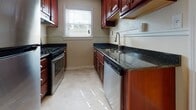 This single-floor home showcases warm hardwood flooring that runs consistently throughout the living areas and bedrooms, lending a cohesive and inviting character to the space. The layout, visible in the floorplan, includes multiple rooms connected by a central corridor, offering a practical and well-organized flow.

The living areas are bright and airy, benefiting from multiple windows dressed with horizontal blinds that allow natural light to filter in and illuminate the polished wood floors. One room features a ceiling fan with integrated lighting, providing both comfort and functionality. Neutral, light-toned walls throughout create a clean, versatile backdrop.

The kitchen is well-appointed with rich cherry wood cabinetry, dark granite countertops, and stainless steel appliances including a gas range and dishwasher. Light tile flooring and a window above the sink add brightness to this functional space.

The bathroom retains a classic retro aesthetic, featuring green ceramic wall tiles accented with black trim, a mosaic tile floor in a dot pattern, a soaking tub, and a window for natural ventilation. The bedrooms are modestly sized, each with hardwood floors and windows offering views of surrounding greenery.