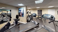 Located in Chattanooga, Tennessee, this fitness center offers a comprehensive workout facility designed to accommodate a wide range of exercise routines. The space is finished with durable rubber flooring throughout, providing both safety and comfort during workouts. A drop ceiling with recessed fluorescent panel lighting ensures consistent, even illumination across the entire floor.

The cardio section is well-stocked with True-brand treadmills and elliptical trainers, offering users high-quality equipment for aerobic exercise. A stationary bike rounds out the cardio offerings. For strength training, the facility includes a full dumbbell rack with an extensive range of weights, a flat weight bench, a barbell with weight plates, and a multi-functional cable machine, catering to both beginners and experienced lifters.

Large wall-mounted mirrors expand the visual space and allow users to monitor their form during exercise. A wall-mounted flat-screen television adds an entertainment option for cardio sessions. Motivational fitness artwork adorns the walls, complementing the energetic teal and neutral beige color scheme that gives the space a vibrant yet professional feel. A posted Fitness Center Rules sign underscores the facility's organized and community-oriented environment. A windowed door provides access to the exterior, allowing a glimpse of natural light into the space.