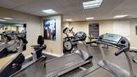 Located in Chattanooga, Tennessee, this fitness center offers a comprehensive workout facility designed to accommodate a wide range of exercise routines. The space is finished with durable rubber flooring throughout, providing both safety and comfort during workouts. A drop ceiling with recessed fluorescent panel lighting ensures consistent, even illumination across the entire floor.

The cardio section is well-stocked with True-brand treadmills and elliptical trainers, offering users high-quality equipment for aerobic exercise. A stationary bike rounds out the cardio offerings. For strength training, the facility includes a full dumbbell rack with an extensive range of weights, a flat weight bench, a barbell with weight plates, and a multi-functional cable machine, catering to both beginners and experienced lifters.

Large wall-mounted mirrors expand the visual space and allow users to monitor their form during exercise. A wall-mounted flat-screen television adds an entertainment option for cardio sessions. Motivational fitness artwork adorns the walls, complementing the energetic teal and neutral beige color scheme that gives the space a vibrant yet professional feel. A posted Fitness Center Rules sign underscores the facility's organized and community-oriented environment. A windowed door provides access to the exterior, allowing a glimpse of natural light into the space.
