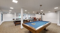 Located in Little Canada, MN, this generously sized indoor recreational facility offers an impressive array of amenities centered around a large, freeform swimming pool with striking blue water. The space spans a single floor and is characterized by soaring ceilings that create an open, airy atmosphere throughout. Tall, angled windows along one wall flood the pool area with abundant natural light while providing views of the surrounding landscape, and multiple glass-panel doors offer direct access to the outdoors.

The pool area is surrounded by a spacious aggregate concrete deck furnished with lounge chairs, seating tables, and dining-style chairs, providing ample room for relaxation. A lower brick wainscoting detail runs along the perimeter walls, accented by decorative artwork that adds a welcoming touch to the space.

Positioned along the poolside wall is an enclosed fitness center, visible through large picture windows. The gym is well-equipped with treadmills, ellipticals, and strength training equipment, offering convenient access directly from the pool deck. An upper-level mezzanine overlooks the pool area, adding architectural depth and vertical interest to the overall design. This facility is well-suited for year-round recreational use, combining aquatic amenities with fitness resources in one cohesive, well-appointed space.