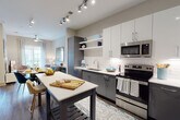 This well-designed single-floor residence offers a cohesive contemporary aesthetic throughout its thoughtfully laid-out living spaces. The kitchen is a standout feature, showcasing a two-tone cabinetry scheme with sleek gray lower cabinets and crisp white upper cabinets, complemented by a classic subway tile backsplash and stainless steel appliances including a range, built-in microwave, dishwasher, and refrigerator. A central island with a white countertop provides additional prep space and casual seating, while track lighting overhead ensures excellent task illumination. The kitchen flows naturally into a comfortable living area, where hardwood-style flooring continues underfoot and a large grid-style mirror enhances the sense of openness. The bedroom is a serene retreat, appointed with plush carpet, a floor-to-ceiling window that draws in natural light, and tasteful wall décor. A dedicated walk-in closet offers adjustable shelving and hanging rails for generous storage. The bathroom maintains the home's clean, modern palette with white quartz countertops, an undermount sink, chrome fixtures, a large vanity mirror, and a tiled shower-tub combination. Throughout, neutral tones and modern finishes create a polished and inviting atmosphere.
