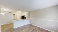 This single-floor unit presents a practical and well-organized layout with a neutral color palette that lends a clean, cohesive feel throughout the space. The living and dining areas are finished with wood-look plank flooring and feature a horizontal chair rail detail along the walls, adding a subtle architectural touch. A front window allows natural light to fill the entry area, while a pendant light fixture illuminates the dining space near the kitchen. A low-profile fireplace platform anchors the living area.

The kitchen is efficiently designed with white shaker-style cabinetry, granite-look countertops, and a peninsula that opens toward the main living space. Black appliances, including a refrigerator and electric range, contrast cleanly against the white cabinetry, and a stainless sink with overhead flush lighting completes the functional workspace.

The bedrooms are carpeted for comfort, each equipped with a ceiling fan. One bedroom features a sliding glass door fitted with vertical blinds, providing access to natural light. A closet is accessible off one of the bedrooms. The bathroom includes a full tub-shower combination with white tile surround and chrome fixtures. Overall, the space maintains a tidy, move-in-ready character throughout.