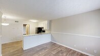 This single-floor unit presents a practical and well-organized layout with a neutral color palette that lends a clean, cohesive feel throughout the space. The living and dining areas are finished with wood-look plank flooring and feature a horizontal chair rail detail along the walls, adding a subtle architectural touch. A front window allows natural light to fill the entry area, while a pendant light fixture illuminates the dining space near the kitchen. A low-profile fireplace platform anchors the living area.

The kitchen is efficiently designed with white shaker-style cabinetry, granite-look countertops, and a peninsula that opens toward the main living space. Black appliances, including a refrigerator and electric range, contrast cleanly against the white cabinetry, and a stainless sink with overhead flush lighting completes the functional workspace.

The bedrooms are carpeted for comfort, each equipped with a ceiling fan. One bedroom features a sliding glass door fitted with vertical blinds, providing access to natural light. A closet is accessible off one of the bedrooms. The bathroom includes a full tub-shower combination with white tile surround and chrome fixtures. Overall, the space maintains a tidy, move-in-ready character throughout.