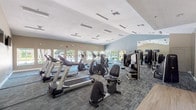 This single-floor fitness center is a comprehensive workout facility designed to support a wide range of exercise routines. The space is outfitted with an impressive selection of cardio equipment, including multiple treadmills, elliptical trainers, and stationary bikes, providing ample options for aerobic training. A dedicated strength training area features several selectorized weight machines suited for full-body conditioning.

The interior design is clean and functional, with patterned carpet tiles in cool blue and grey tones covering the floor throughout. Warm beige walls contrast with a striking accent wall painted in a soft blue-grey, lending the space a calm, motivating atmosphere. Vaulted ceilings contribute to an open, airy feel, and the room is brightly lit by rows of recessed linear fluorescent fixtures.

Large windows along multiple walls invite abundant natural light and offer views of an outdoor pool area and lush greenery, creating an energizing environment for workouts. Full-length mirrors are strategically positioned to enhance the sense of space and allow users to monitor their form. Wall-mounted flat-screen televisions provide entertainment during cardio sessions. The layout is thoughtfully organized to ensure clear circulation between equipment zones, making the facility both practical and comfortable for users.