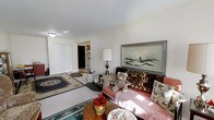 This single-floor space presents a warm and lived-in living room as its centerpiece. The room is finished with wall-to-wall carpeting and features neutral white walls that lend a clean, bright backdrop to the interior. Natural light filters into the space, complementing a ceiling-mounted light fixture overhead.

A set of built-in bi-fold closet doors lines one wall, offering generous storage within the living area. The room accommodates a cushioned sofa adorned with decorative pillows and a classic landscape painting in a silver frame mounted above it, adding an artistic focal point to the space. Layered area rugs in traditional patterns add texture and define the seating zone.

A wooden side table holds a vintage-style table lamp, contributing to the warm ambiance of the room. A round coffee table anchors the seating arrangement, while decorative accents throughout reflect a cozy, personal aesthetic.

An open passage leads toward what appears to be additional rooms, including a glimpse of a tiled area suggesting a kitchen or entryway nearby. Overall, the space offers a comfortable and functional single-level layout with a classic, homey character.