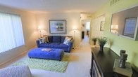 This single-story space centers around a welcoming living room finished with wall-to-wall carpeting in a warm neutral tone. The room accommodates a full-sized blue sofa dressed with decorative pillows and paired with a matching tufted ottoman, creating a comfortable and cohesive seating arrangement. A shag area rug in light green anchors the seating area, adding texture and a pop of color to the neutral palette. Framed artwork on the wall and a floor lamp contribute to the inviting ambiance. A dark media console sits along one wall, offering both storage and display space, accompanied by potted greenery that adds a natural accent. The walls are painted in soft, warm tones, with one accent wall in a muted green, providing subtle visual contrast. An open corridor visible from the living room leads to additional spaces, suggesting a flowing, connected layout. Natural light filters through a window visible in the adjoining room, brightening the overall atmosphere. The space presents a functional and comfortable layout well-suited for everyday living.