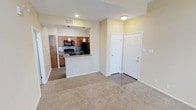 This single-floor residence offers a thoughtfully designed open-concept layout that seamlessly connects the living area and kitchen. The living space features wall-to-wall carpeting in neutral tones, creating a warm and comfortable atmosphere, while recessed lighting and a flush-mount ceiling fixture provide consistent illumination throughout.

The kitchen is a functional centerpiece of the home, anchored by a peninsula with a dark stone countertop that doubles as a breakfast bar. Rich warm-toned wood cabinetry lines the walls, complemented by a classic tile backsplash that adds visual texture. The kitchen is fully equipped with black appliances, including a refrigerator, stove, and microwave, and is accented by striking red pendant lights that hang above the peninsula, adding a bold pop of color to the space.

The transition between the carpeted living area and the tiled kitchen floor creates a clear, natural division between the two spaces. A paneled interior door near the kitchen leads to an additional room, offering flexible use of space. The soft, neutral wall tones throughout lend the unit a clean, move-in-ready feel. Overall, the space balances practicality with inviting design details, making it a comfortable and functional home.
