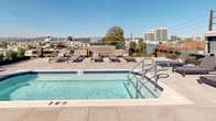Located in the heart of West Los Angeles at 11311 La Grange, this rooftop amenity deck offers an exceptional outdoor experience with striking panoramic views of the surrounding cityscape. The space is defined by its clean, modern aesthetic, featuring crisp white walls, large-format tile flooring, and sleek glass railings that preserve unobstructed sightlines to the Los Angeles skyline and beyond.

At the center of the rooftop is a sparkling pool framed by tiled edging and equipped with stainless steel entry rails, surrounded by a generous sun deck furnished with contemporary chaise lounges — ideal for relaxing under the abundant Southern California sun. A dedicated bar area features a long, built-in counter finished in a neutral tone, lined with natural wood stools that lend a warm contrast to the otherwise modern palette. Potted plants and decorative greenery add organic accents throughout the space.

Architectural details include frosted glass privacy screens, open staircase access, and thoughtful lighting elements that suggest versatility for both daytime lounging and evening gatherings. The expansive open sky and far-reaching urban views make this rooftop a standout amenity, blending sophisticated design with a relaxed, resort-inspired atmosphere.