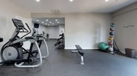 The fitness center at Steeplechase Apartment Homes, located at 3700 W Steeplechase Way in Williamsburg, Virginia, is a well-appointed, single-floor facility designed to support a comprehensive workout routine. The space is accessed through a tiled entry corridor branded with the Steeplechase Apartment Homes logo, providing a clean and welcoming transition into the facility.

The main workout area features durable rubber flooring throughout, offering both safety and comfort during exercise. Recessed lighting illuminates the space evenly, creating a bright and functional environment. A large floor-to-ceiling mirror spans one wall, visually expanding the room and allowing residents to monitor their form during workouts.

The cardio section is equipped with multiple treadmills, elliptical machines, and additional aerobic equipment, while the strength training area includes a multi-station cable machine, adjustable benches, and free weights. A wall-mounted television provides entertainment during cardio sessions.

A secondary room offers additional open floor space ideal for stretching or flexibility work, complete with a stability ball, a wall-mounted exercise guide chart, and a bench for convenience. Natural light filters in through a window in the main workout area, adding to the overall energetic ambiance. The facility is well-ventilated with ceiling-mounted HVAC units and is accessible via a glass-paneled interior door.