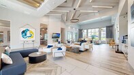 Located at 3200 Champion Ring Rd in Fort Myers, Florida, this single-floor amenity clubhouse offers a generously sized, thoughtfully designed gathering space for residents and guests. The grand main lounge immediately impresses with soaring ceilings, coffered beam detailing, and elegant pendant and chandelier lighting. Large floor-to-ceiling windows and glass doors flood the interior with natural light and provide views of the outdoor pool area. Light wood-grain tile flooring in a herringbone pattern anchors the main seating area, which features contemporary white lounge chairs, plush sofas, and a central fireplace with a wall-mounted display above it. Adjacent to the lounge, a coffee bar with dark countertops and high stools provides a casual gathering spot. The game room offers a lively retreat, complete with a billiards table, comfortable seating, a wall-mounted television, and stylish pendant lighting above an exposed wood beam ceiling. A dedicated conference room features a large table, ergonomic seating, and glass-paneled doors that connect to additional breakout spaces. A business center workstation with built-in countertops, pendant lighting, and a textured stone accent wall supports productivity. A package room with shelving and large windows rounds out the functional amenities of this well-appointed community facility.